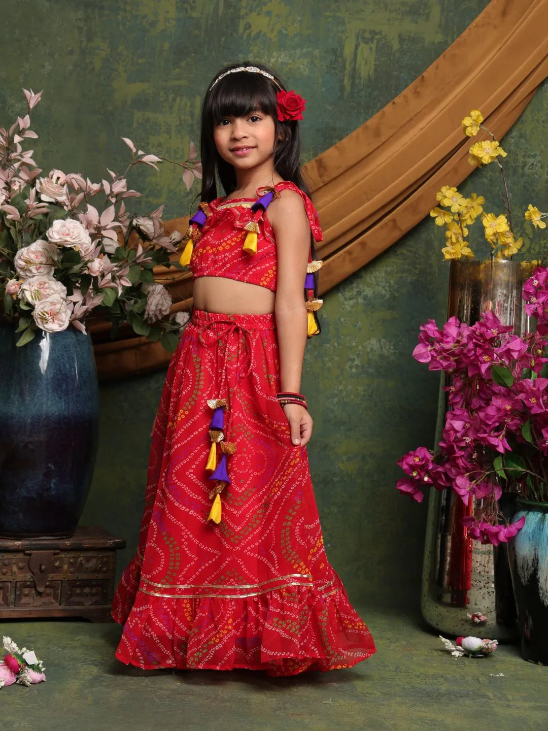 Young girl in a red traditional outfit with colorful tassels standing in front of floral decorations.