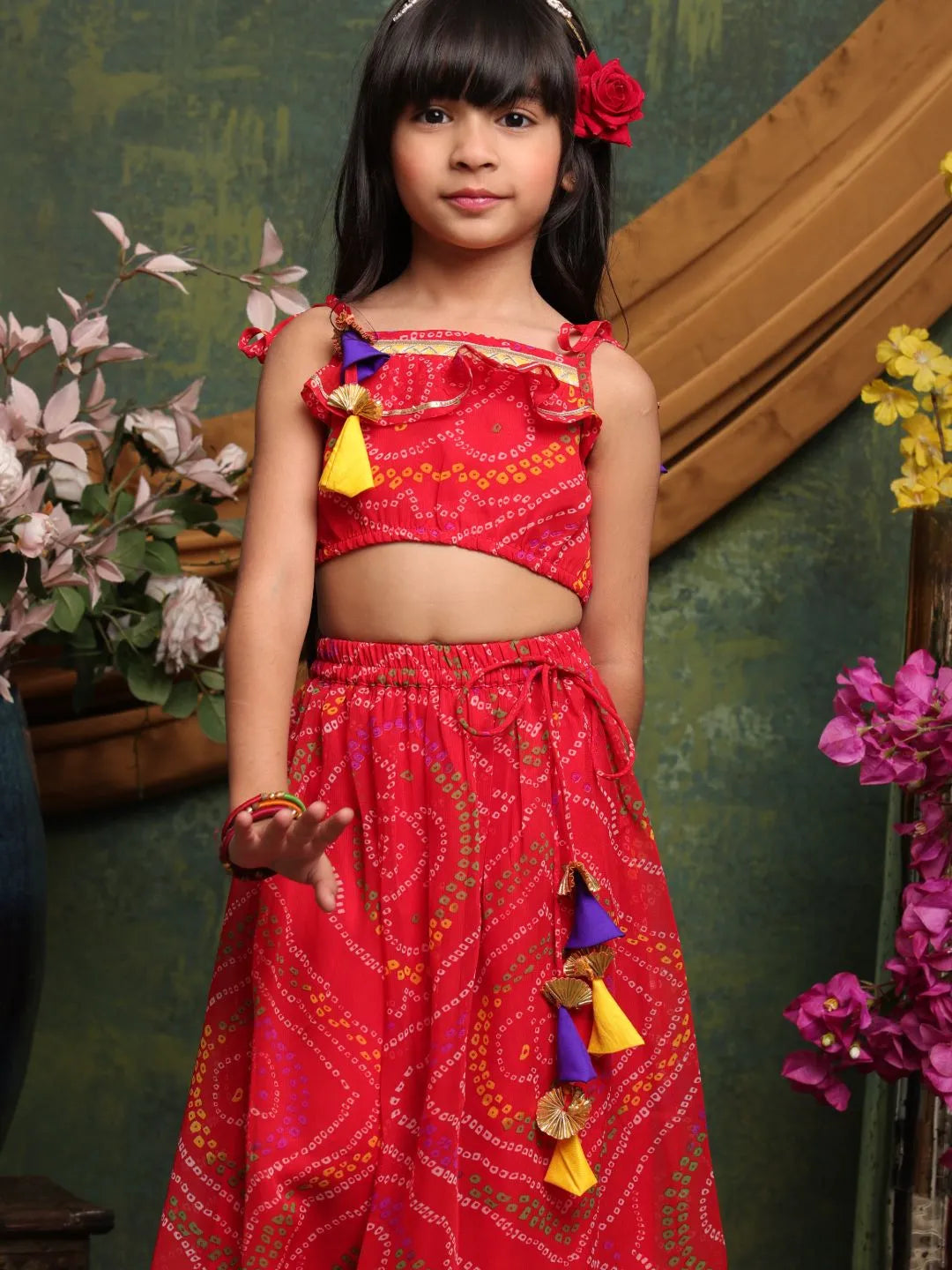 Young girl in a red traditional outfit with colorful accents standing in front of a decorative wall.