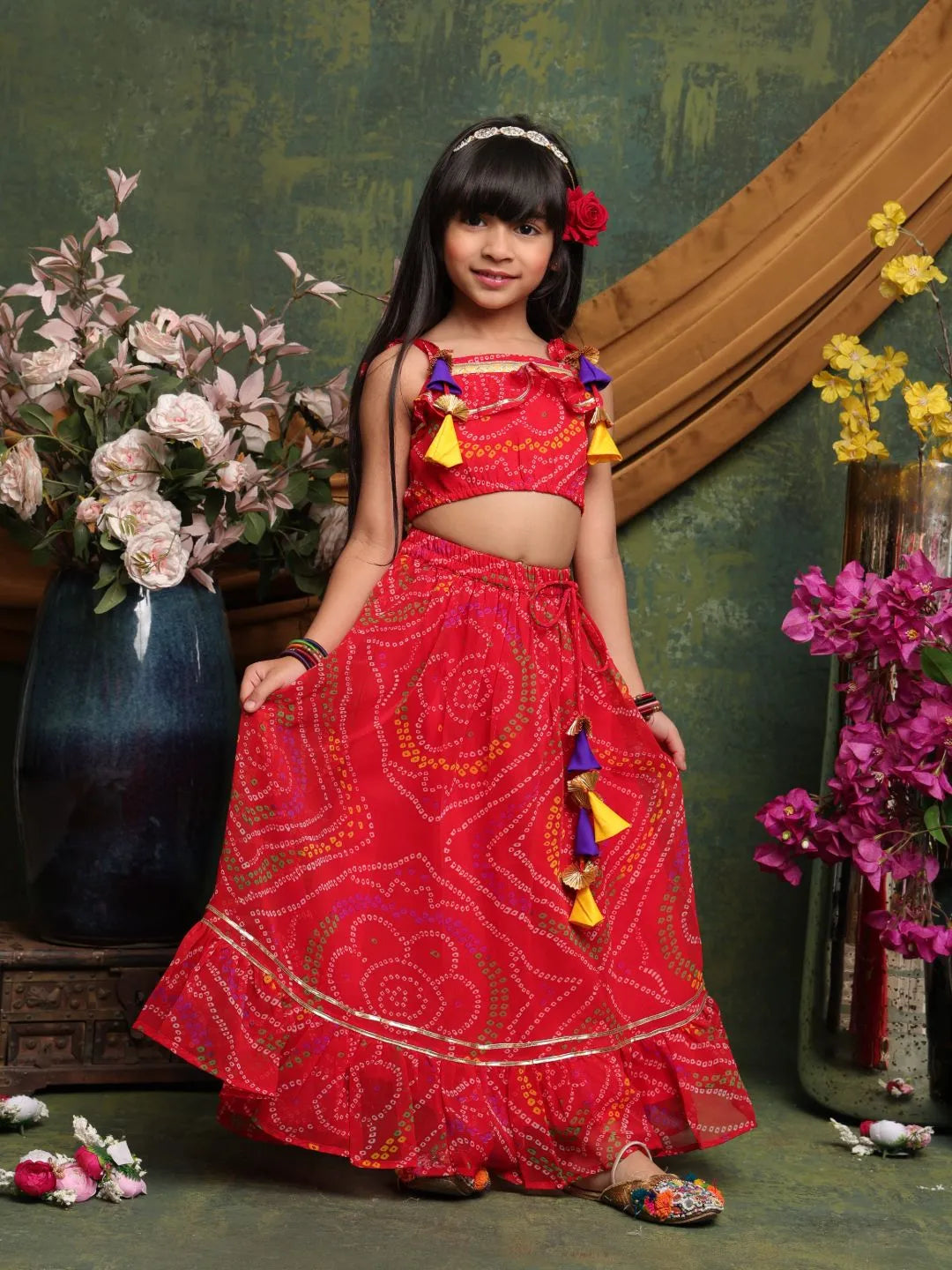 Young girl in a red traditional outfit with colorful accents standing in front of floral decorations.