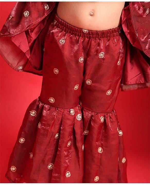 A close-up of a child wearing a red traditional outfit with gold embroidered motifs, featuring a flared skirt and matching draped sleeves, set against a solid red background.