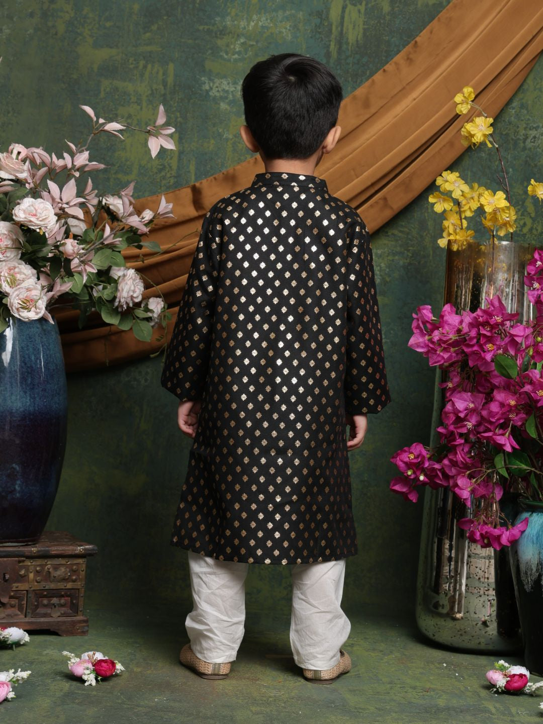 Child wearing a black and white patterned kurta with white pants standing in front of decorative flowers and vases.