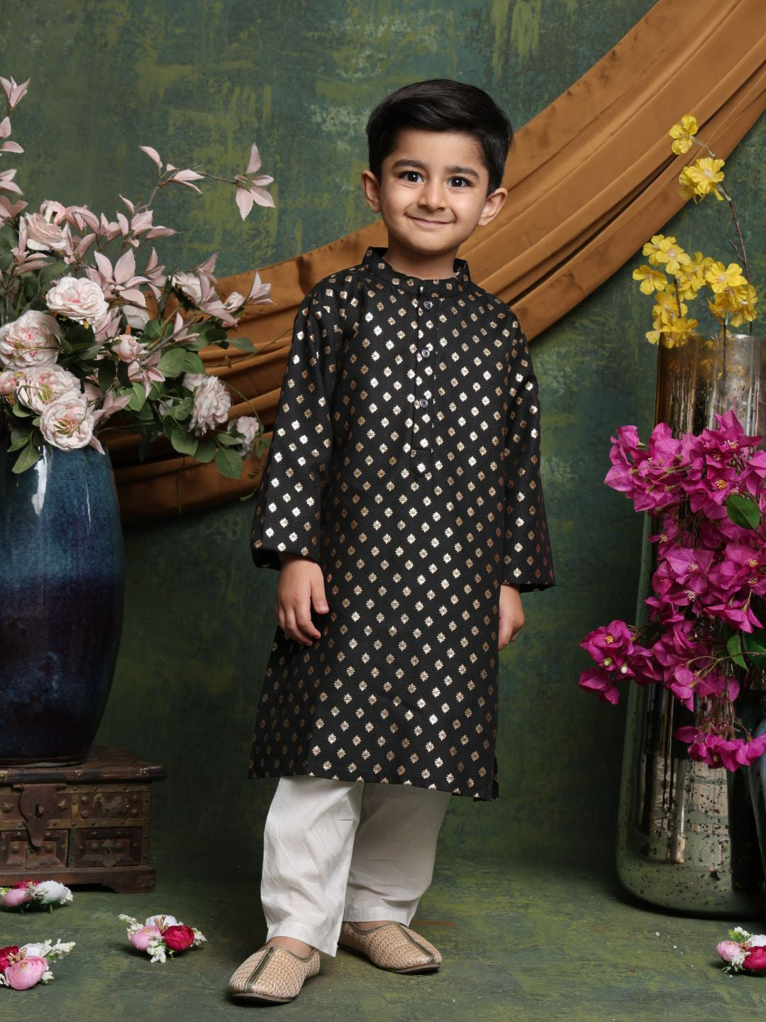 Child wearing a black kurta with white patterns standing in front of decorative flowers and a green wall.