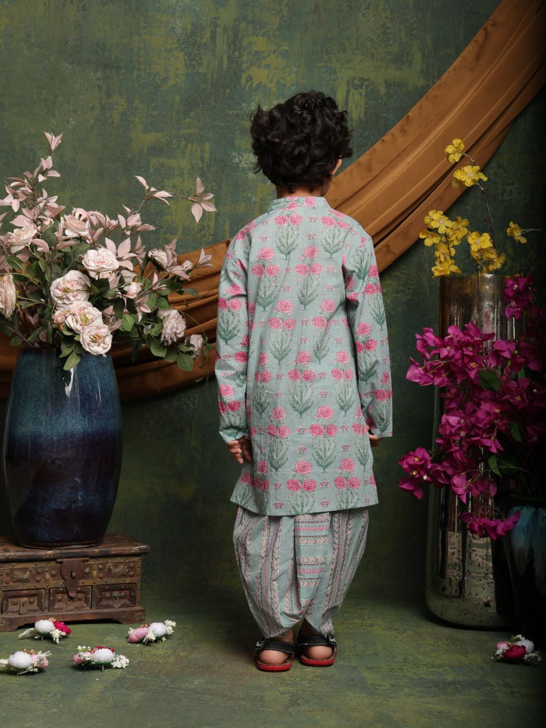 Person wearing a floral outfit standing in front of a decorative mirror with flowers on a green wall.