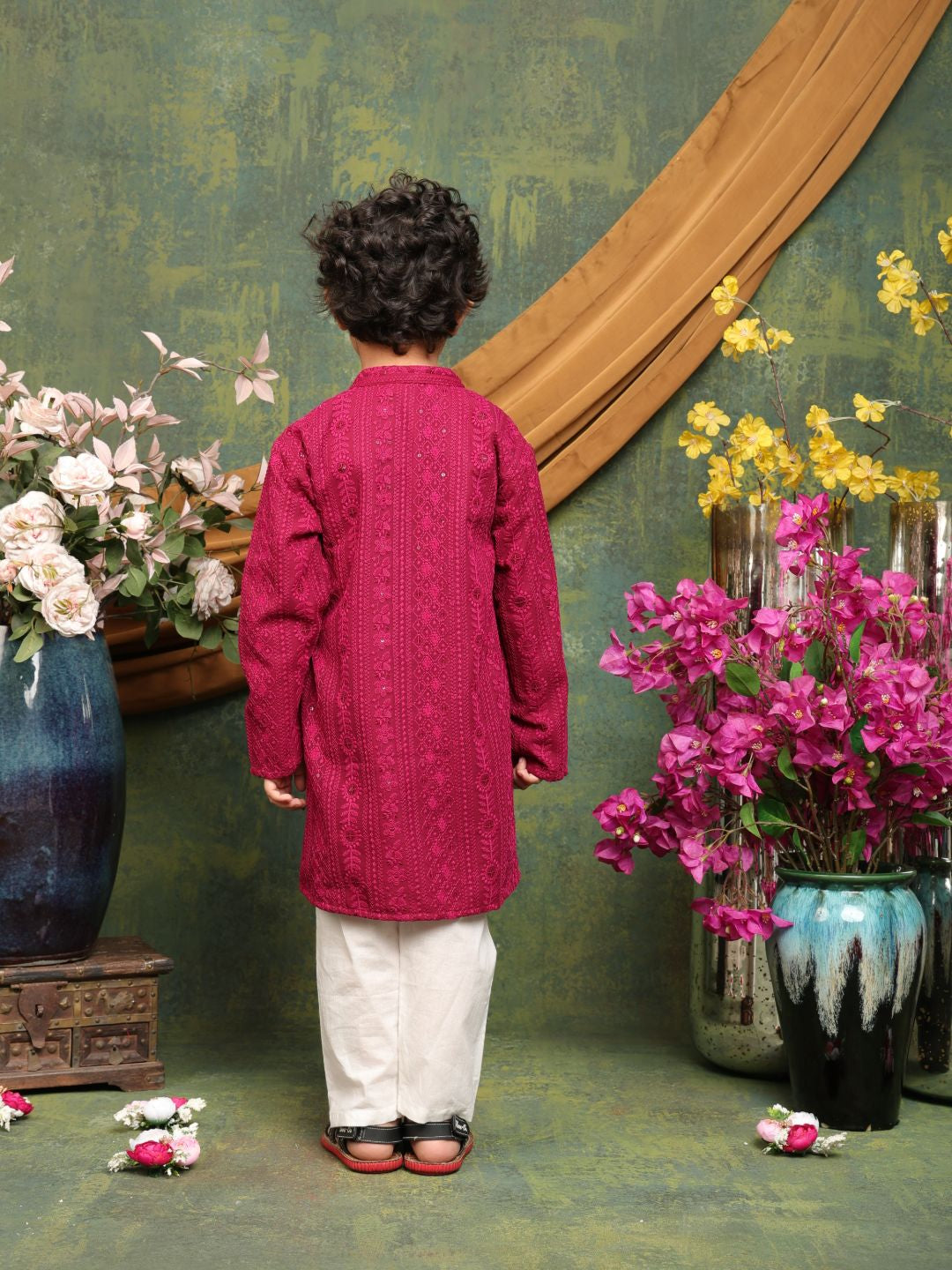 Child wearing a red kurta with white pants standing in front of floral arrangements.