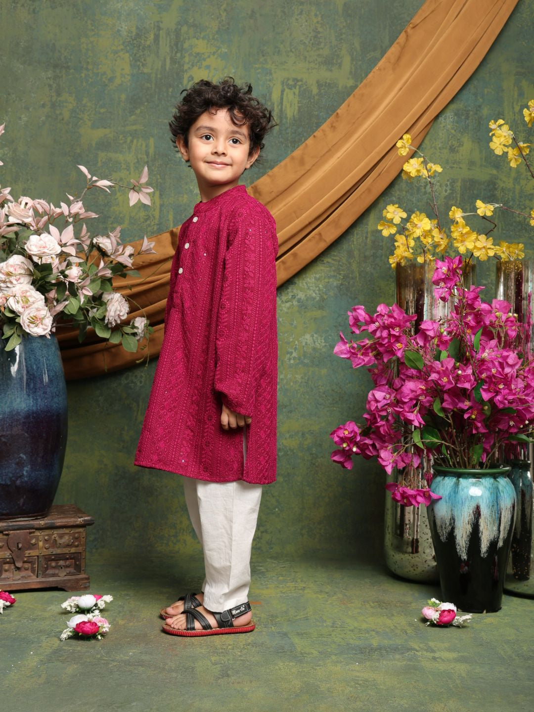 Child in a red kurta with white pants standing in a decorative setting with flowers and vases.