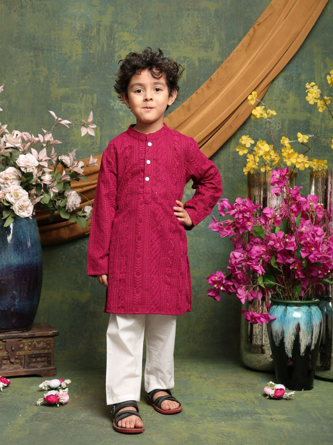 Child wearing a pink kurta with white pants standing in front of floral decorations.