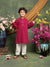 Child wearing a red kurta with white pants standing in front of floral decorations.