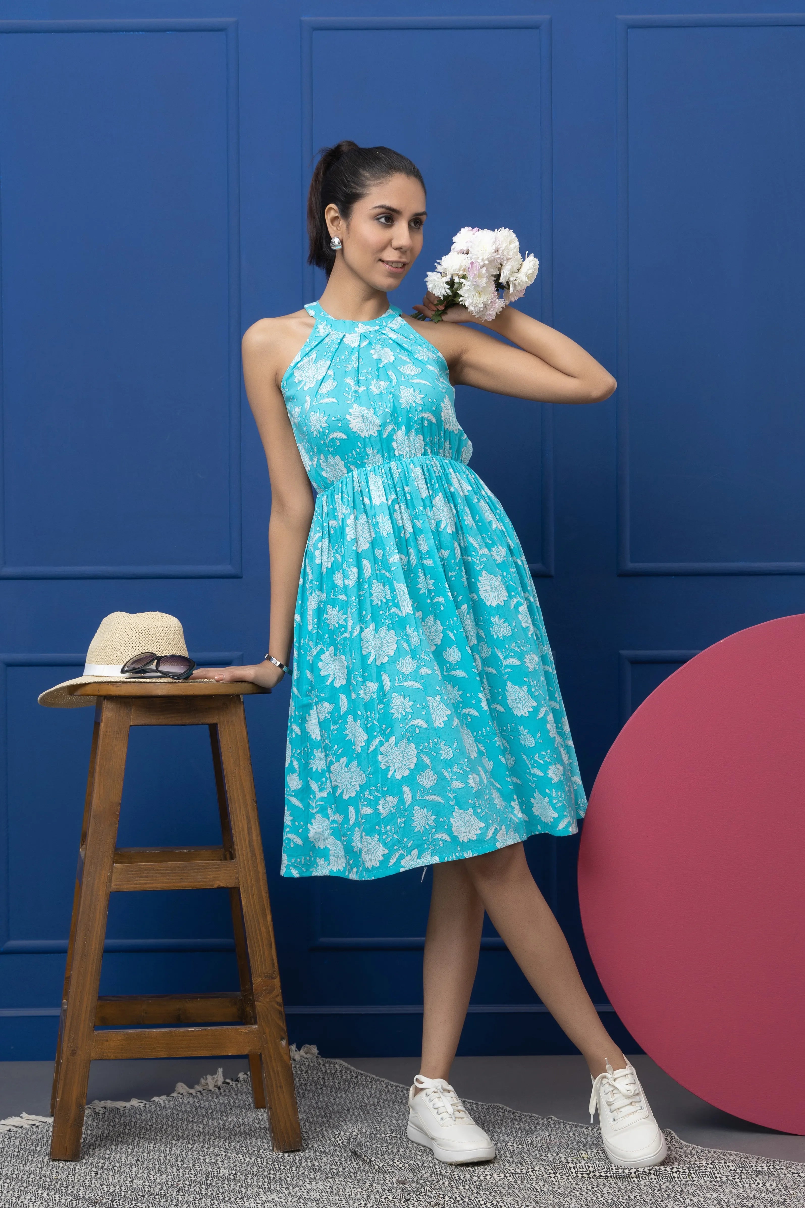 Woman in a blue floral dress posing against a blue wall with a wooden stool and pink object.