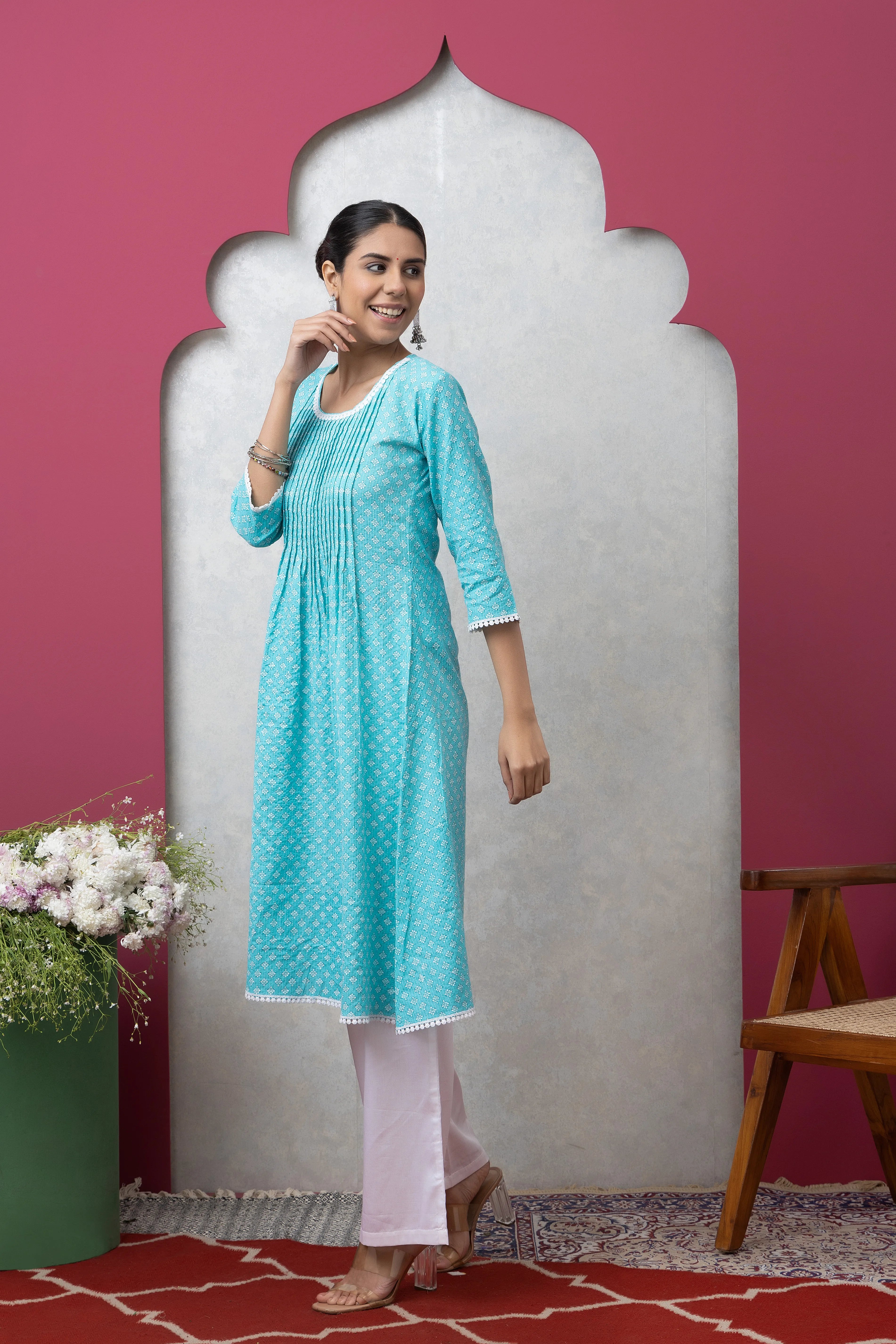 Woman wearing a turquoise kurta with white pants against a pink wall.