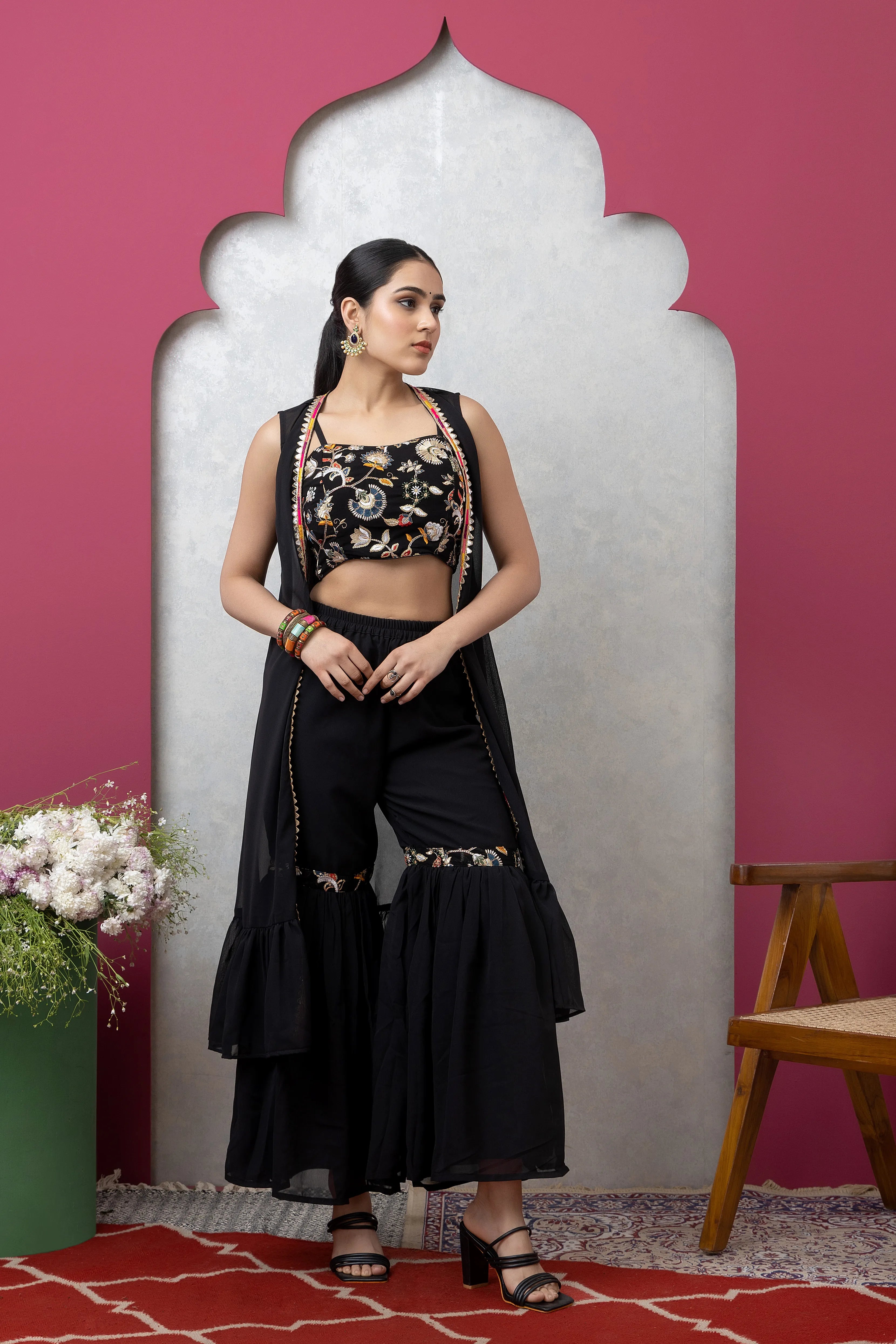 Woman in black floral top and black pants posing against a pink wall with a decorative mirror.