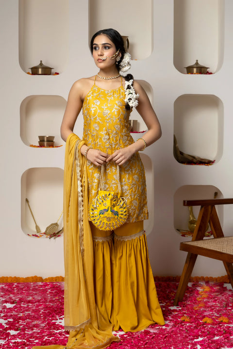Woman in a yellow traditional outfit with floral patterns standing against a decorative wall.