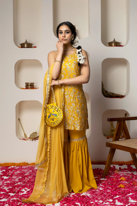 Woman in a yellow traditional outfit with floral patterns standing against a decorative wall.