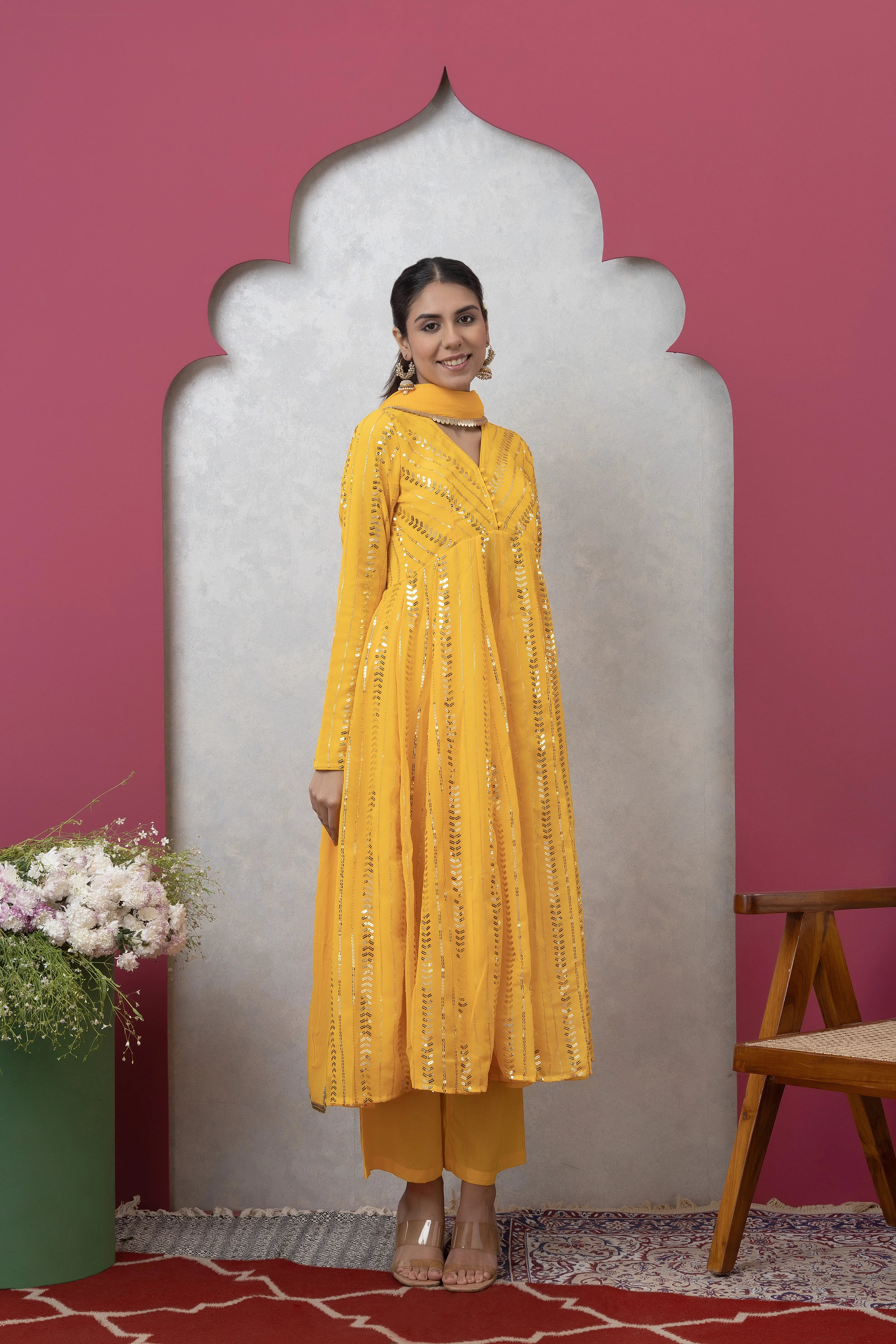Woman wearing a yellow embroidered outfit standing against a pink wall with decorative elements.