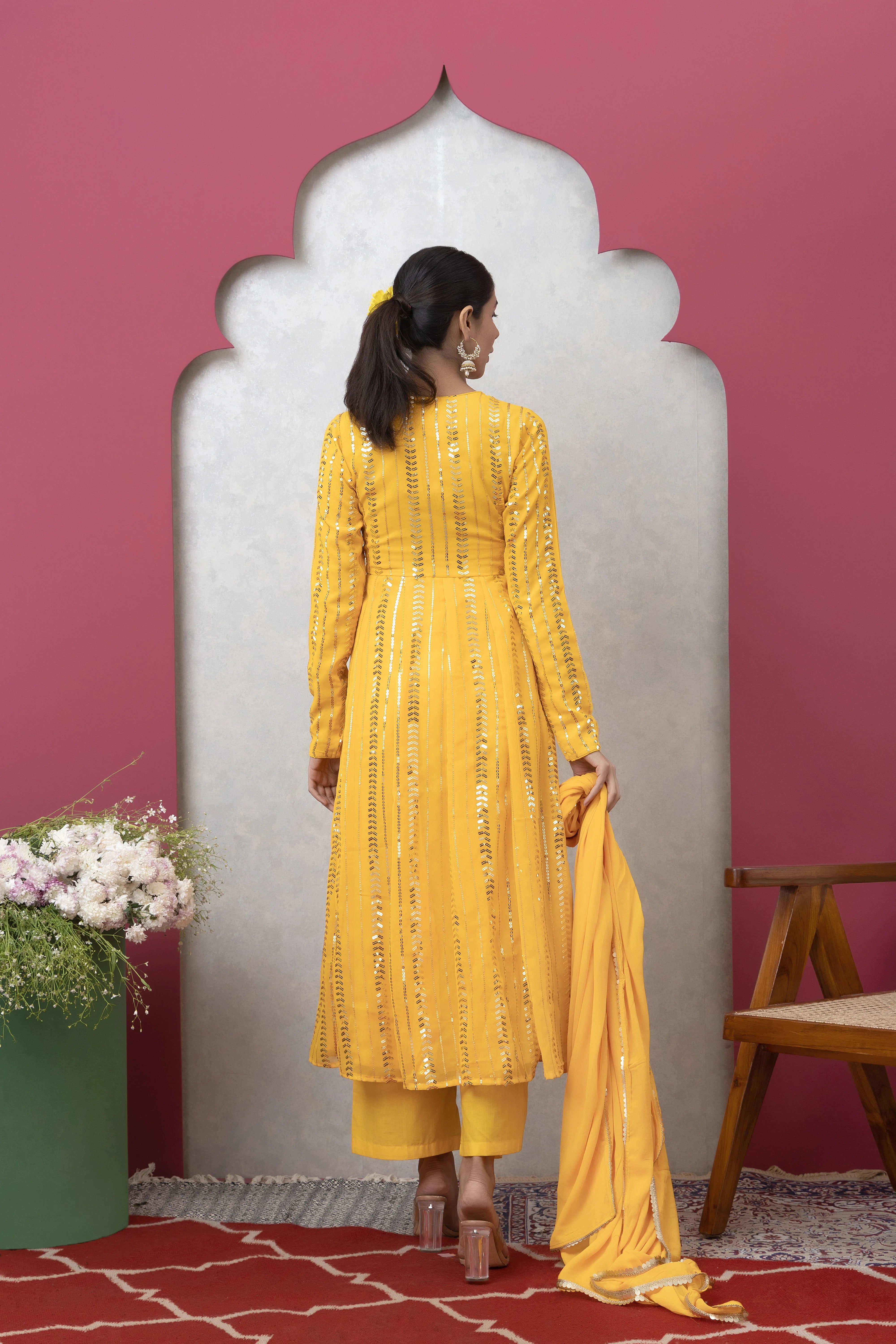 Woman in a yellow traditional outfit standing in front of a decorative archway with a pink wall.