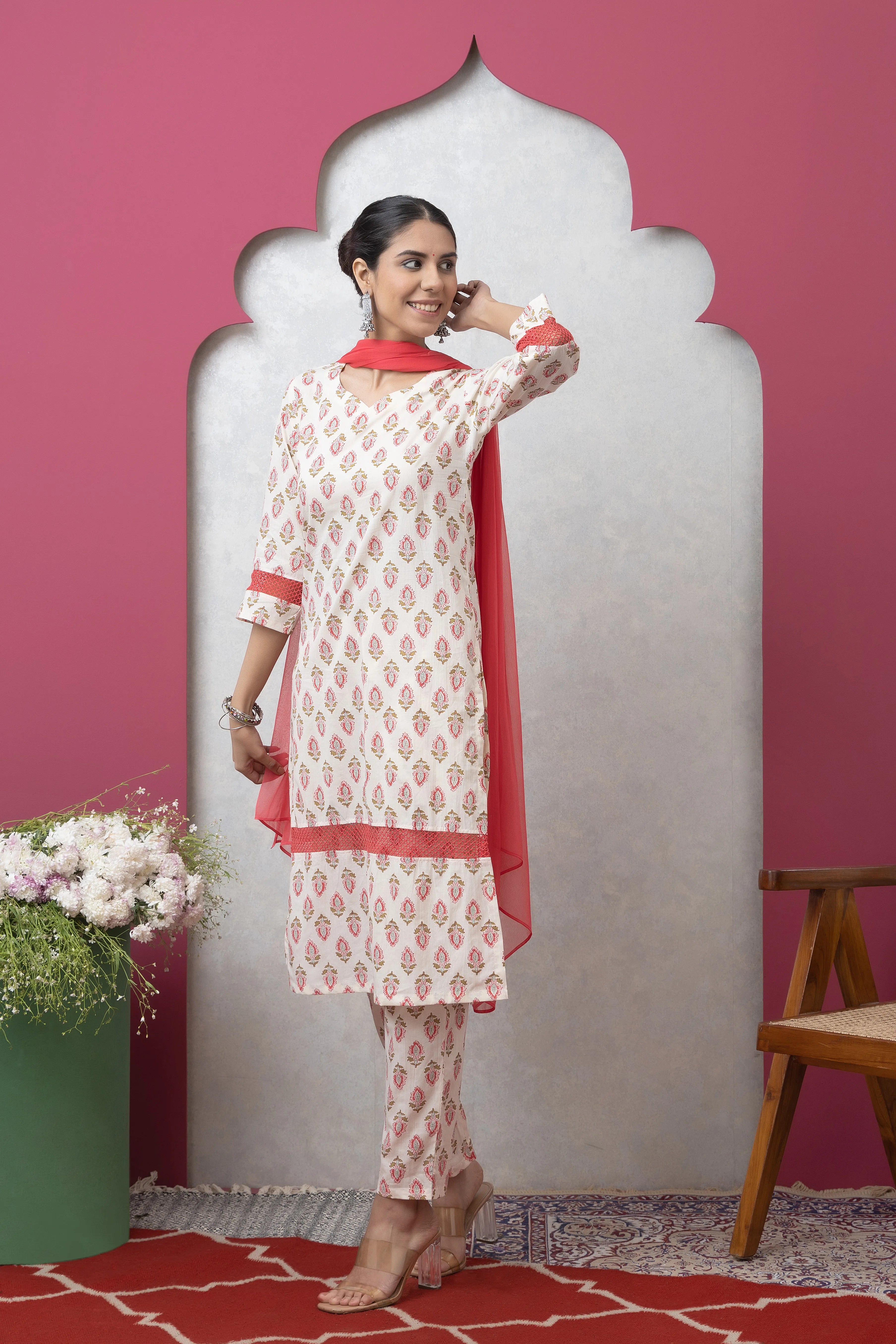 Woman in a white and red traditional outfit standing against a pink wall with a decorative arch.