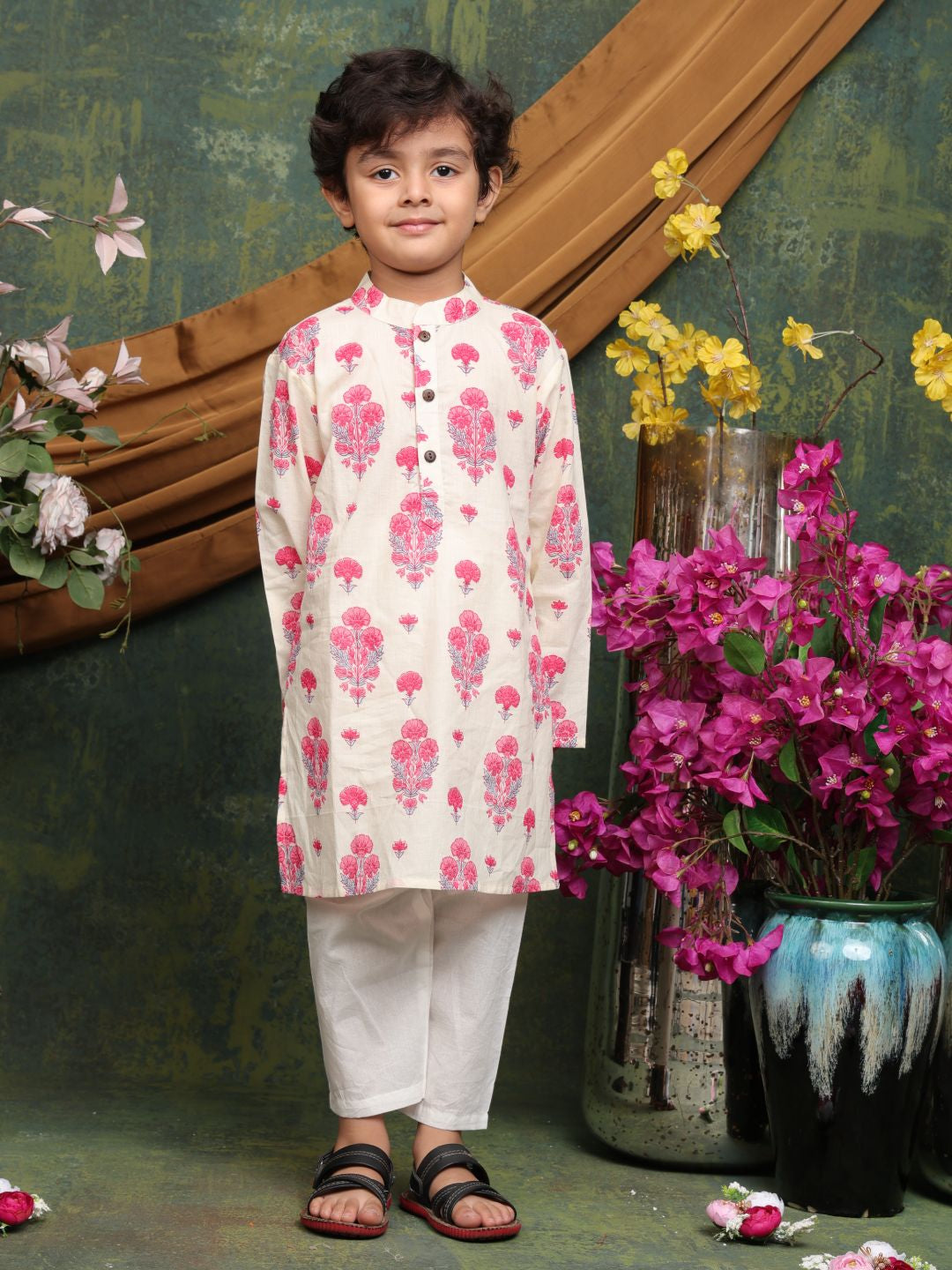 Child wearing a floral kurta with white pants standing in front of decorative flowers and plants.