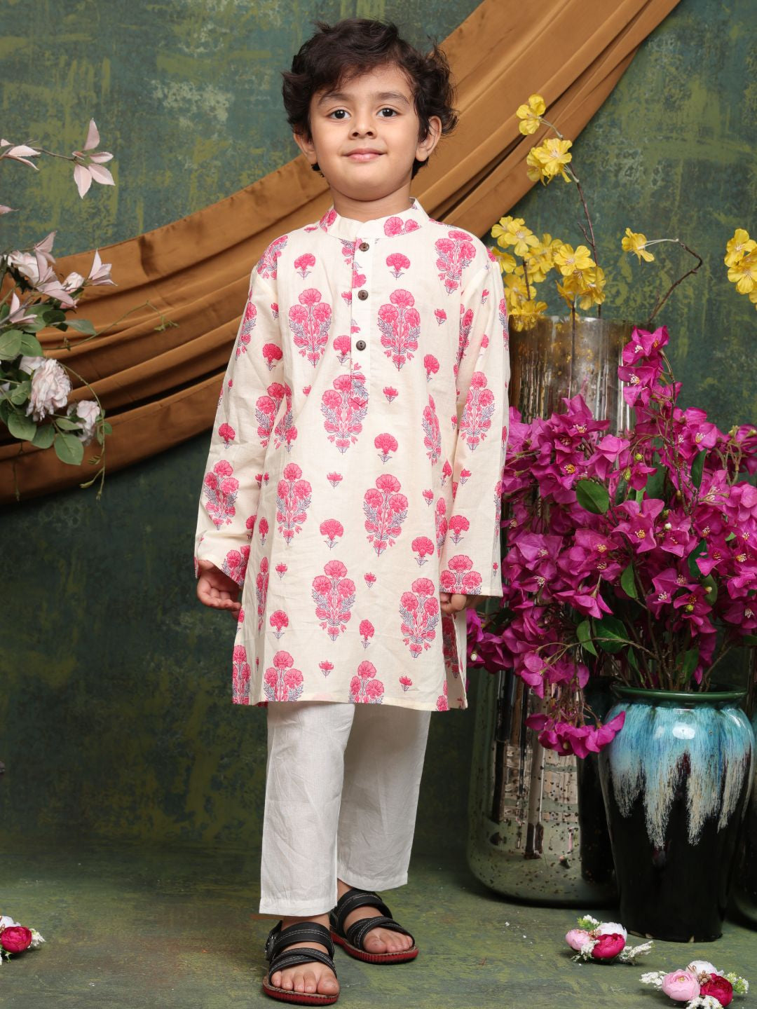 Child wearing a floral kurta with white pants standing in front of decorative flowers and vases.