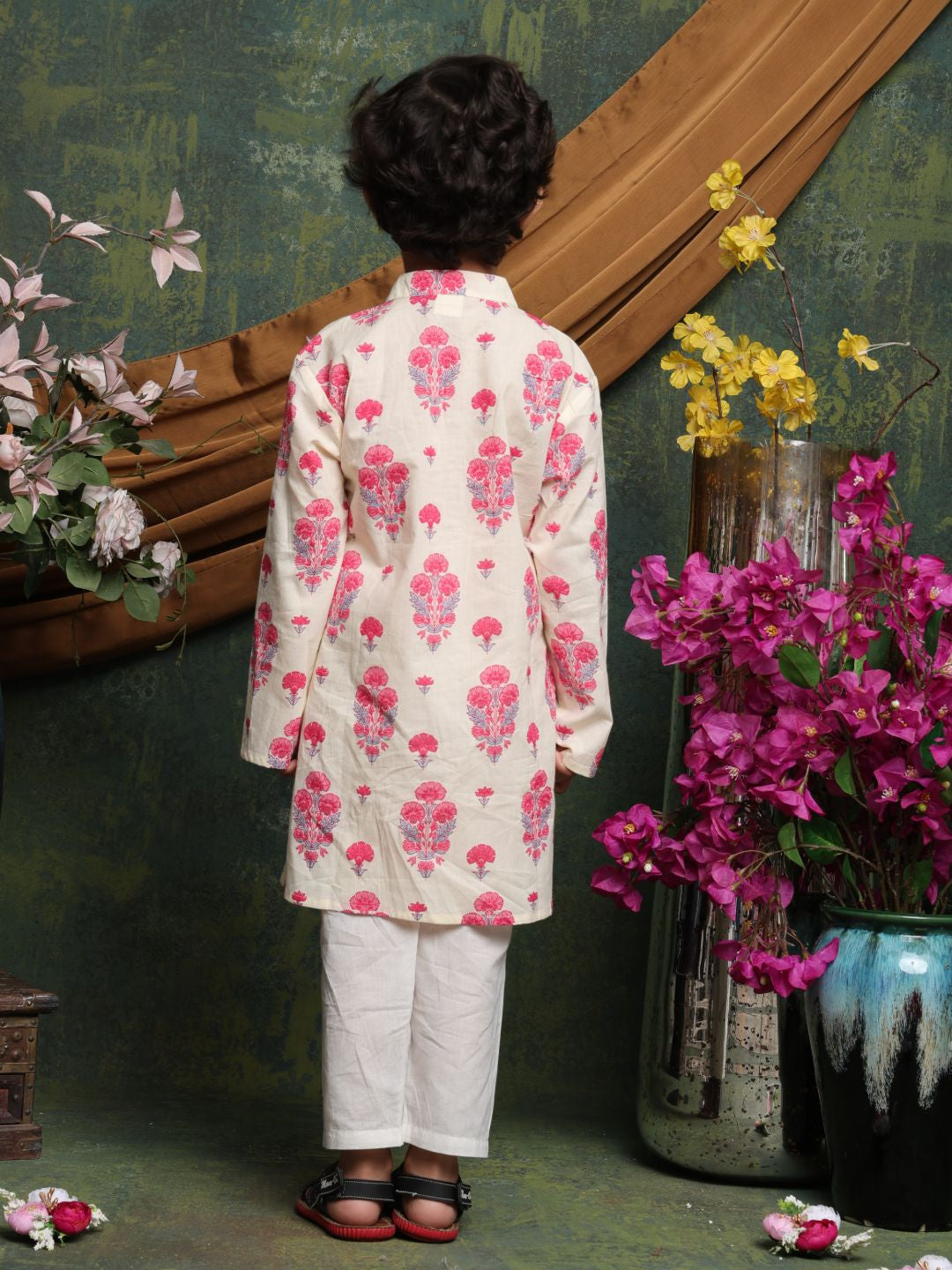Person wearing a floral kurta with white pants standing in front of a decorative wall with flowers.