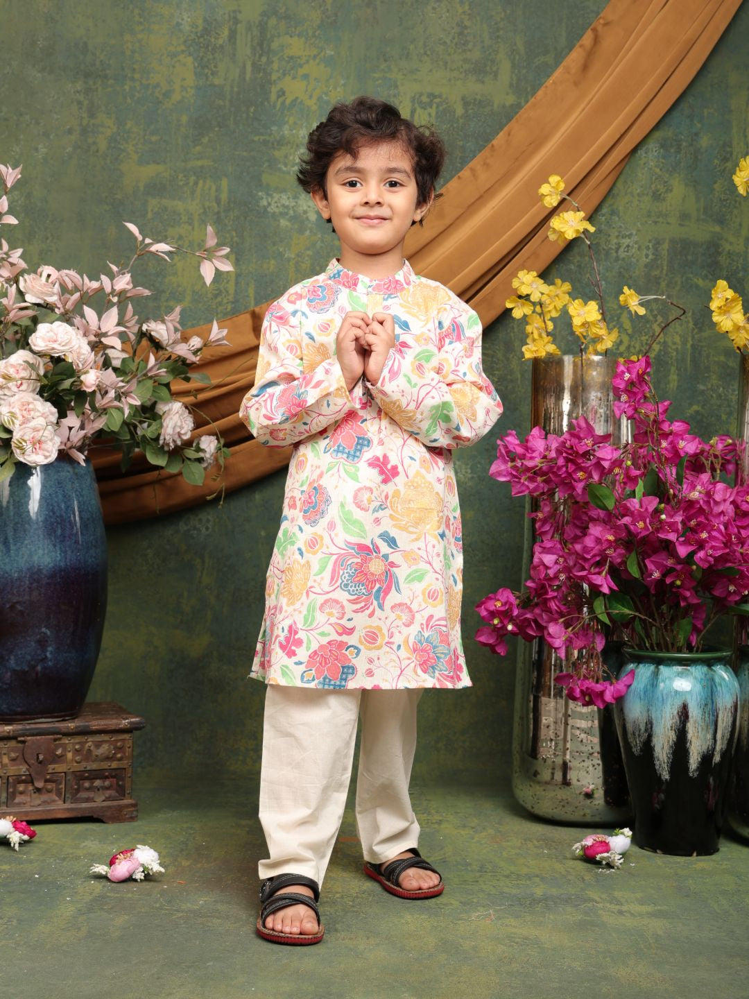 Child in a floral kurta with white pants standing in front of decorative vases and flowers.