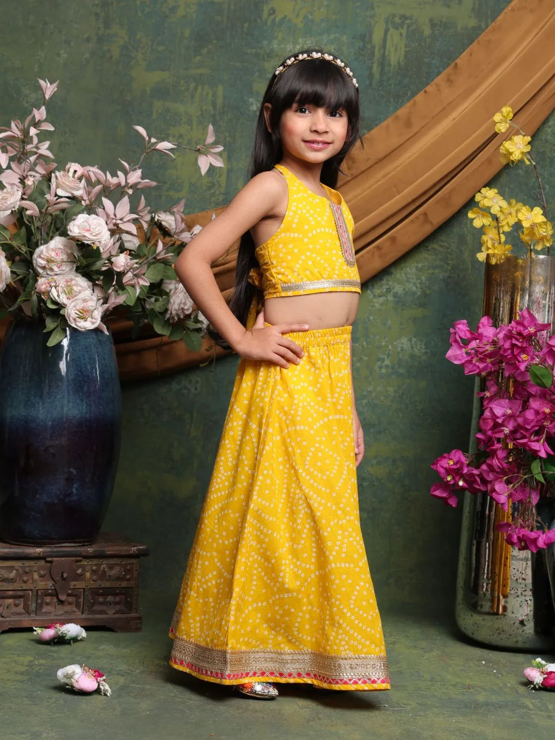Young girl in a yellow traditional outfit standing in front of decorative flowers and a mirror.