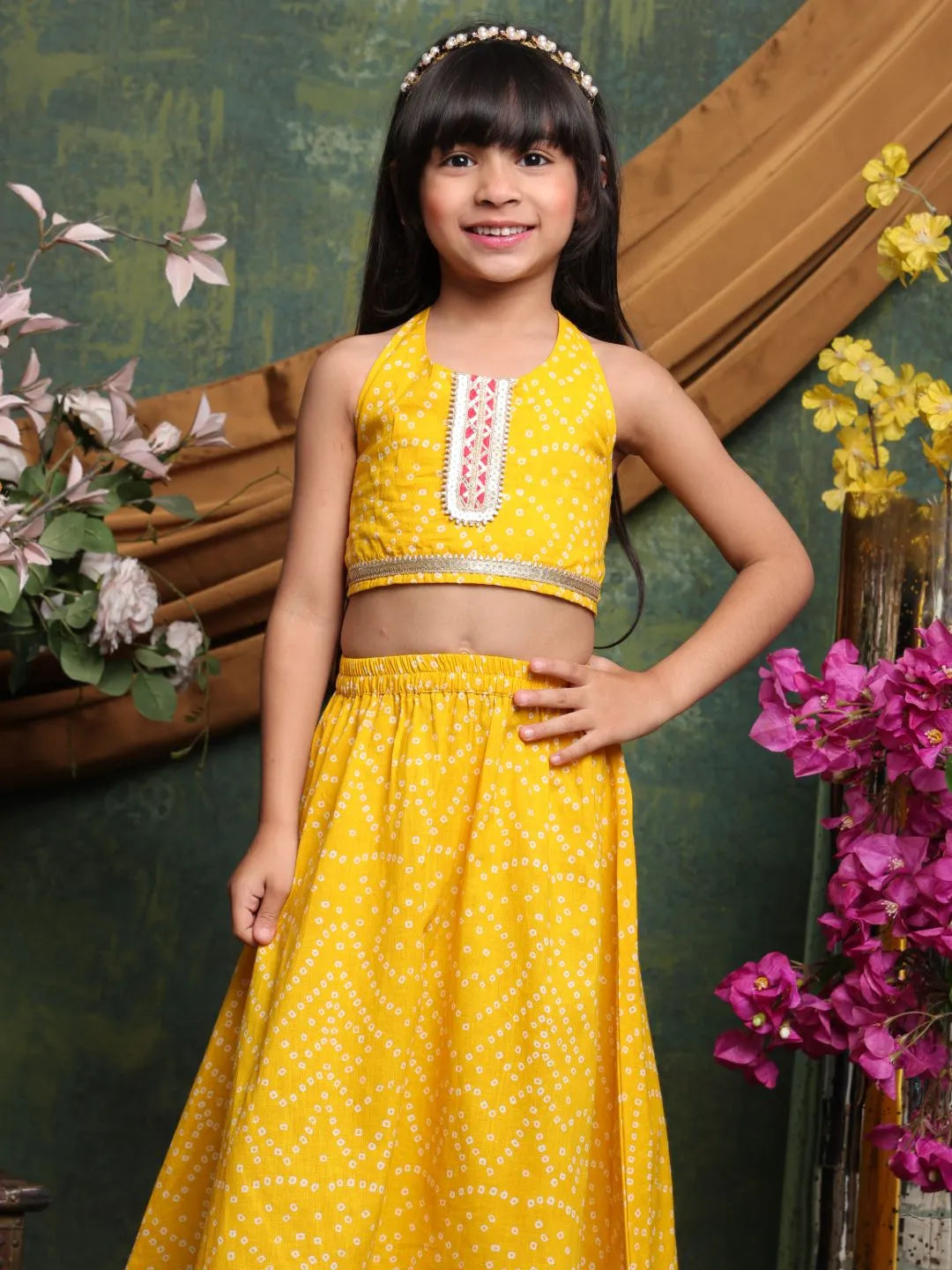 Young girl in a yellow traditional outfit standing in front of floral decorations.