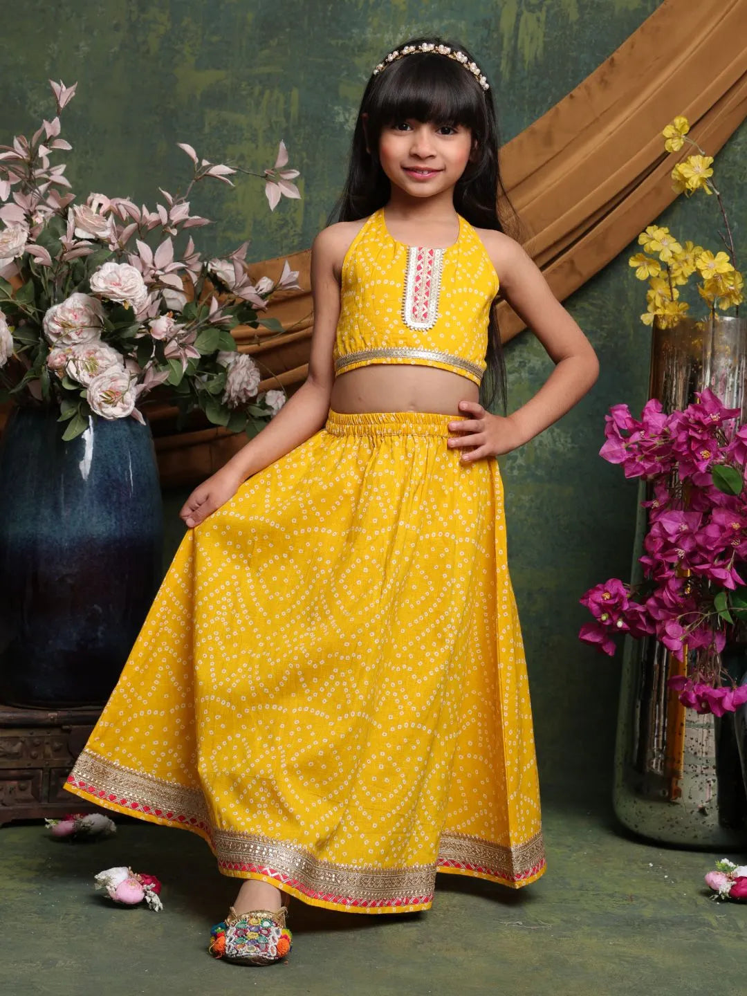 Young girl in a yellow traditional outfit standing in front of floral decorations.
