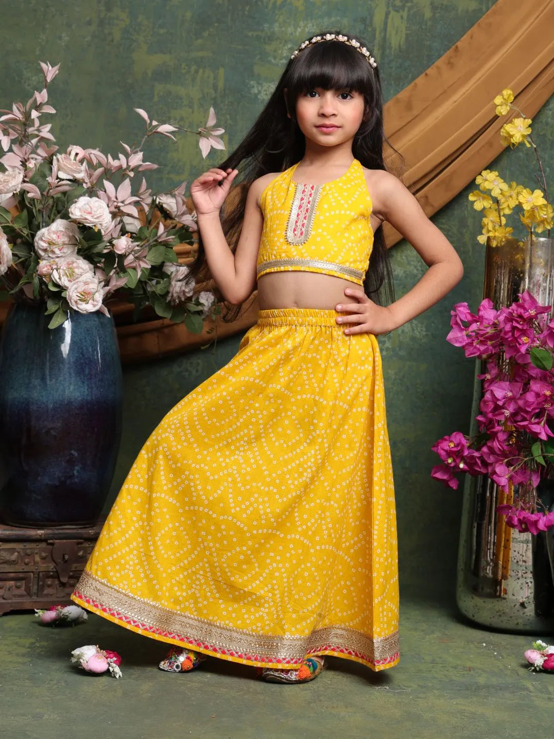 Young girl in a yellow traditional outfit standing in front of floral decorations.