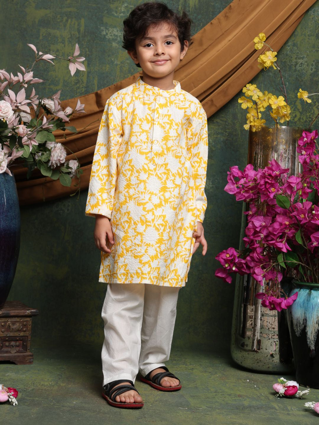 Child wearing a yellow and white kurta with white pants standing in front of floral decorations.