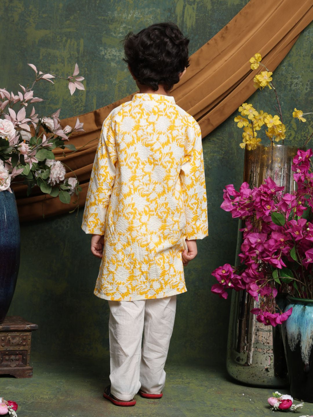 Child wearing a yellow and white traditional outfit with floral patterns, standing in front of a decorative wall with flowers.