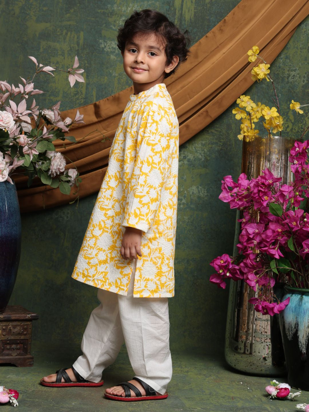 Child wearing a yellow and white traditional outfit with floral decorations and flowers in the background.