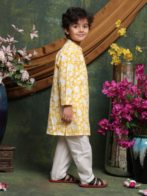 Child wearing a yellow and white floral kurta with white pants, standing in front of decorative flowers.