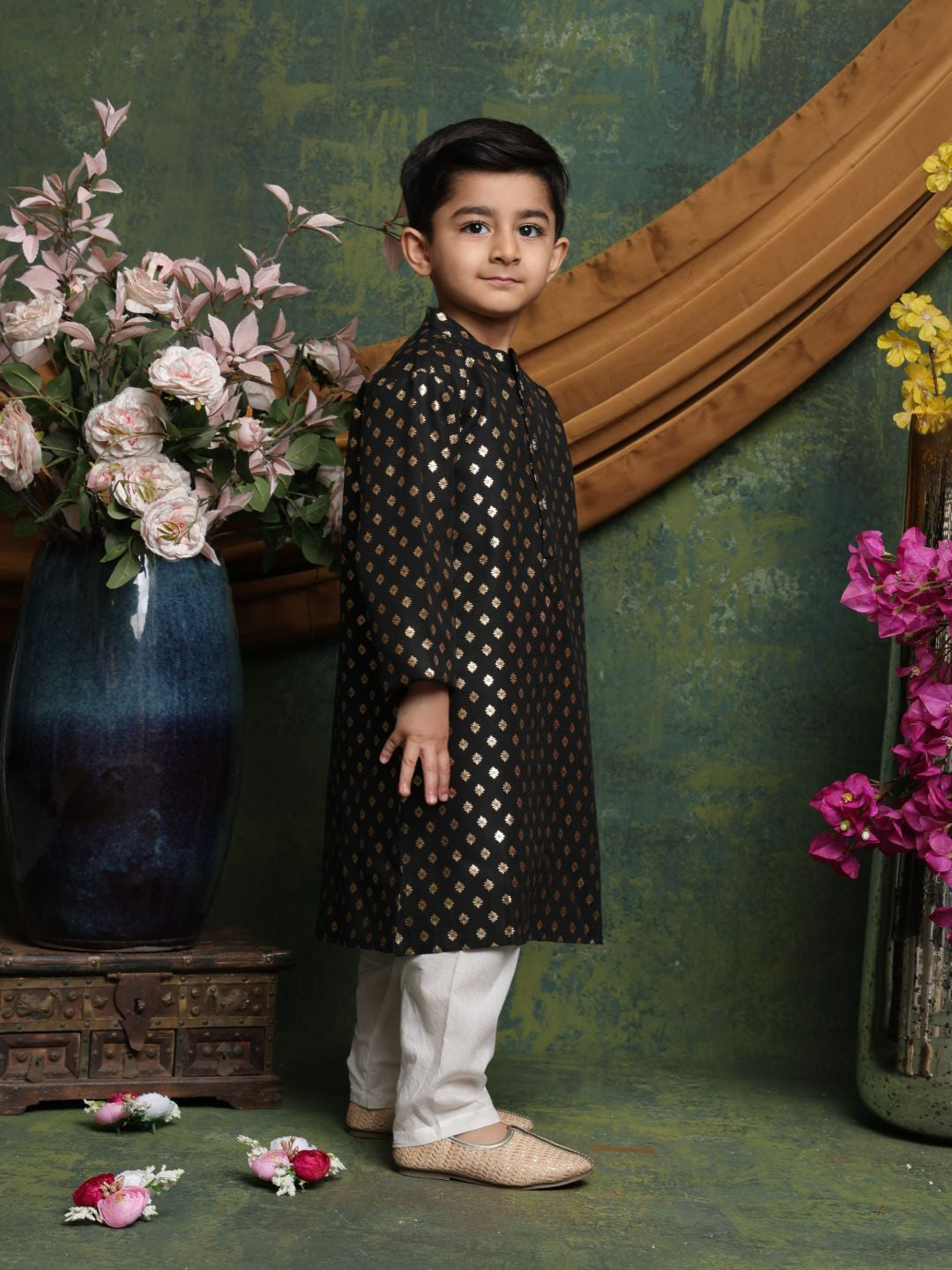 Young boy in a traditional outfit standing in front of decorative flowers and a mirror.