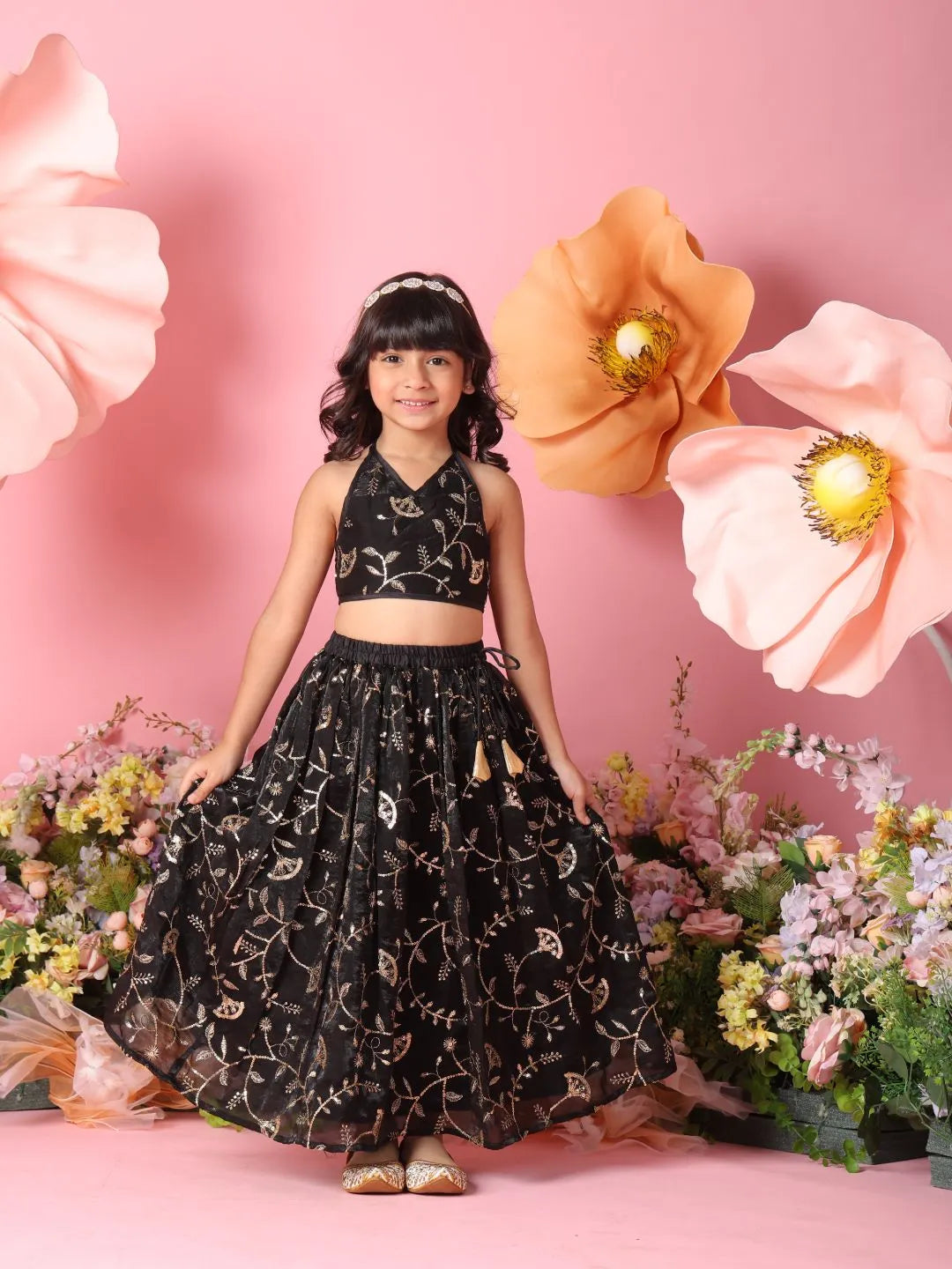Young girl in a black embroidered outfit standing against a pink background with large flowers.