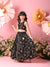 Young girl in a black floral dress standing against a pink background with large flowers.