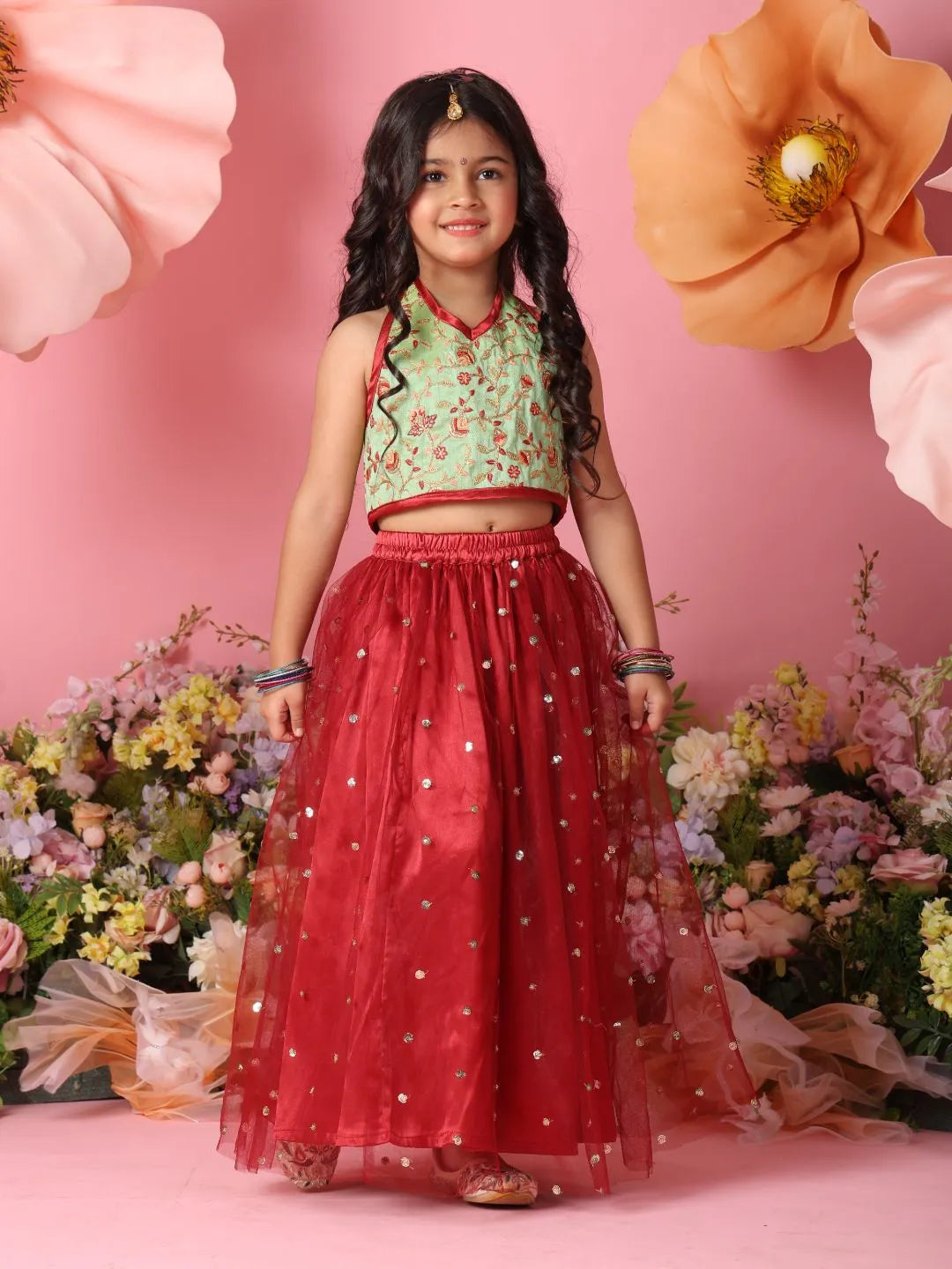 Young girl in a green top and red skirt standing against a pink background with flowers.