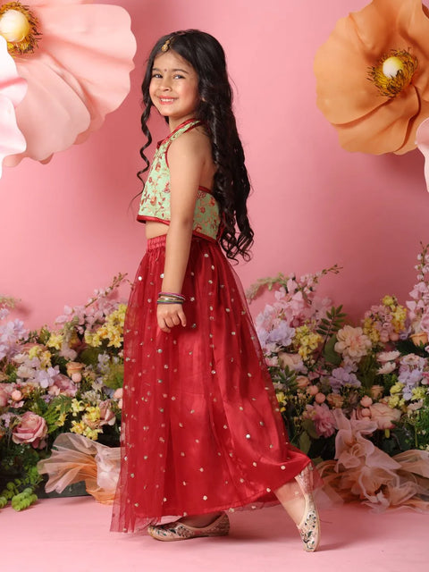 Young girl in a green top and red skirt standing against a pink background with flowers.