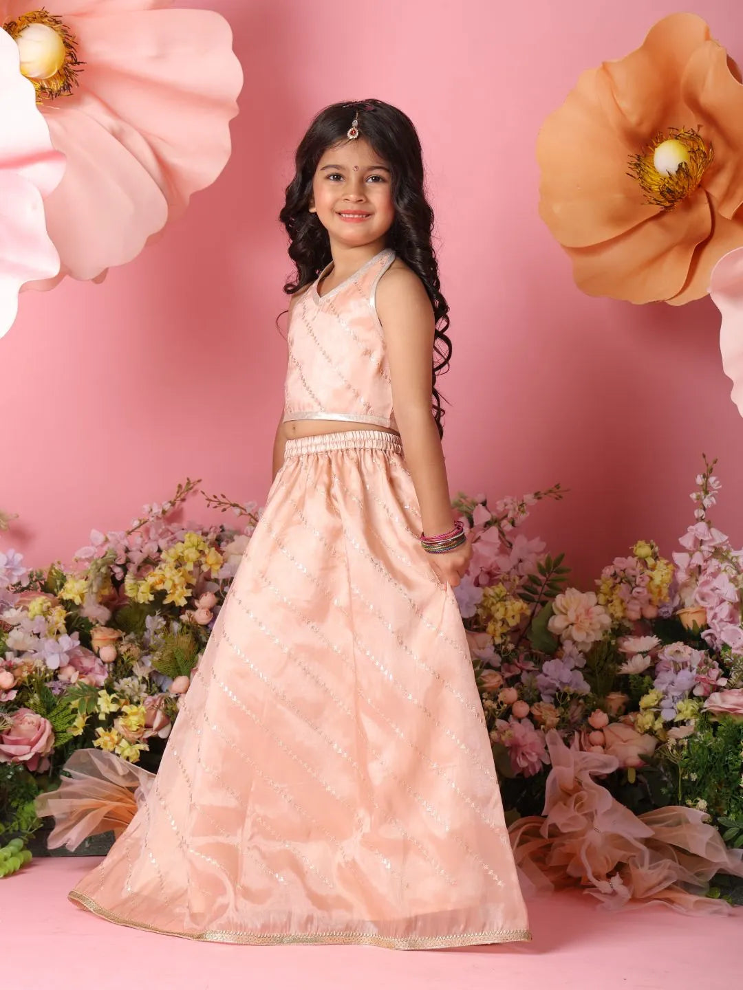 Young girl in a peach traditional outfit standing against a pink background with flowers and decorative elements.