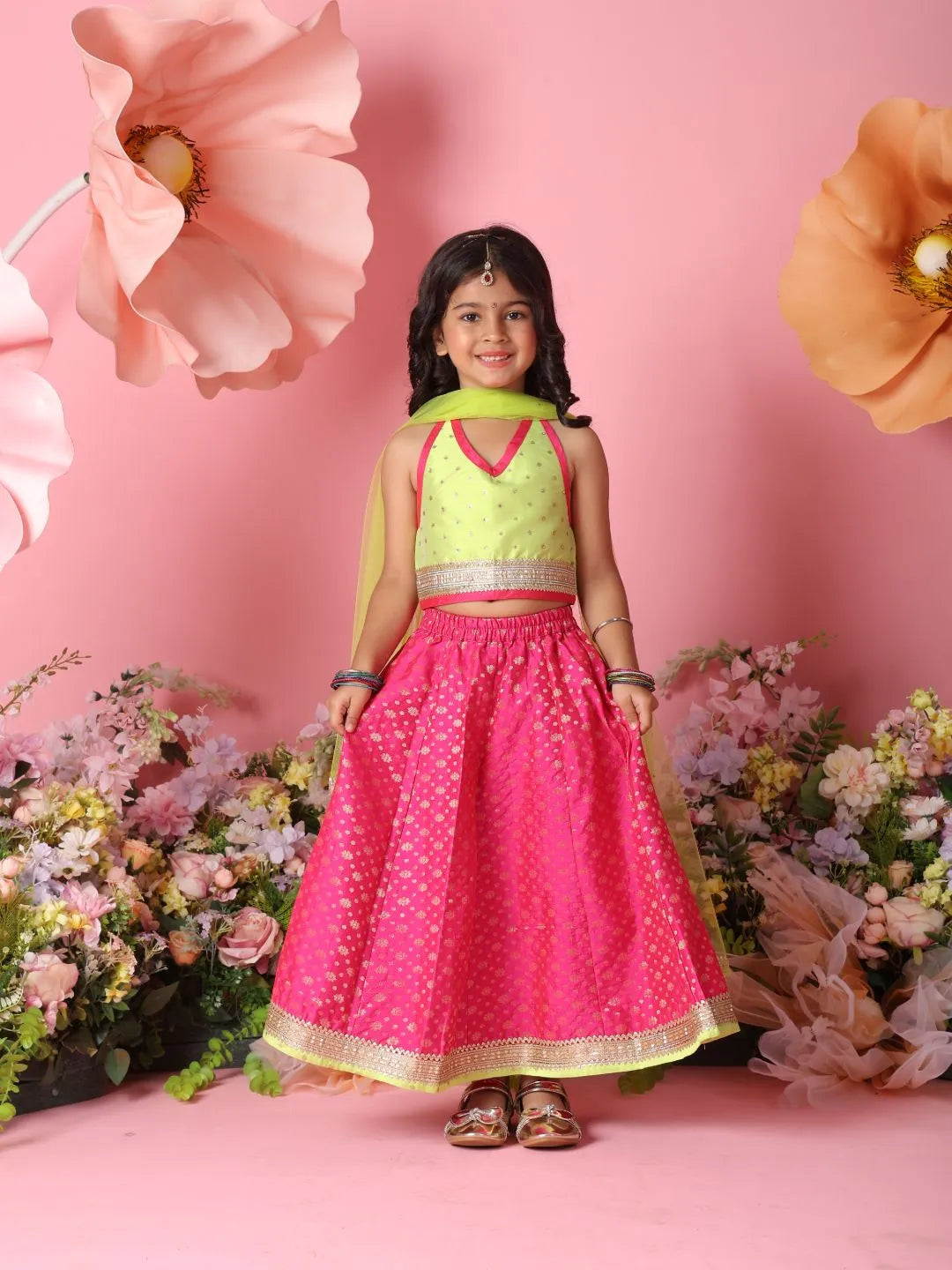 Young girl in a pink and green traditional outfit standing against a pink background with flowers.