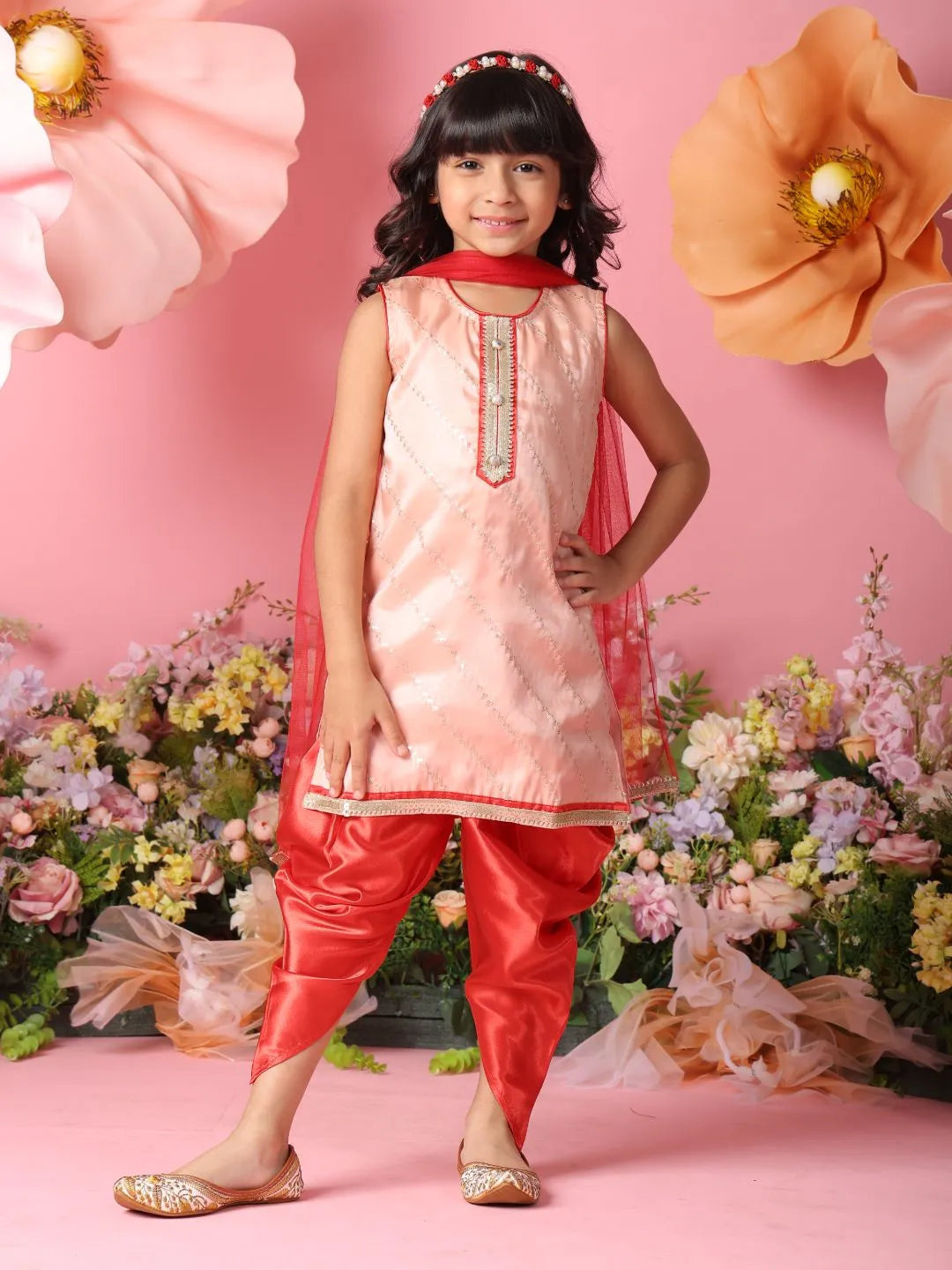Young girl in a pink and red traditional outfit standing against a pink background with flowers.