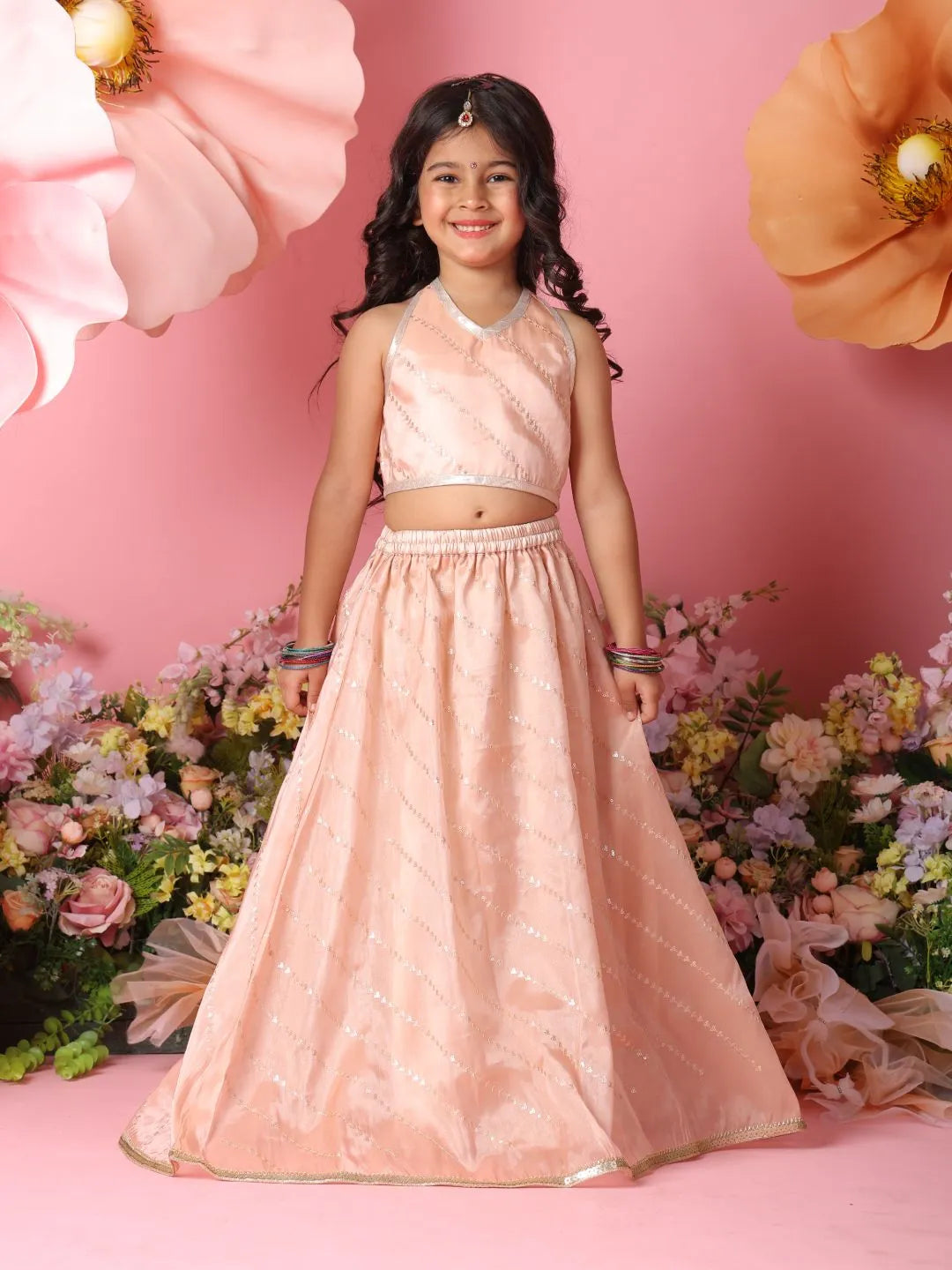 Young girl in a pink traditional outfit standing against a floral backdrop with a pink background.