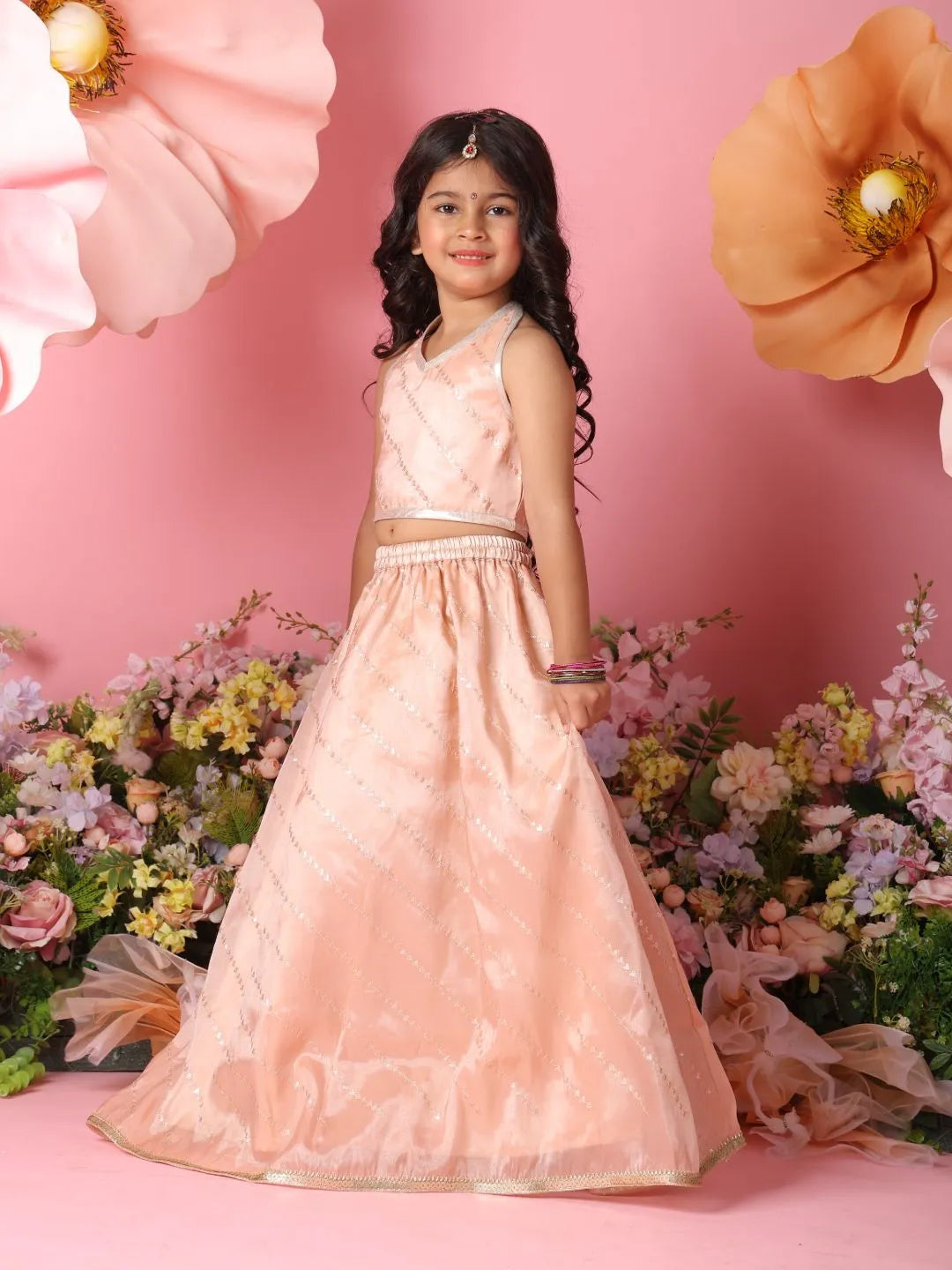 Young girl in a pink traditional outfit standing against a pink background with flowers.