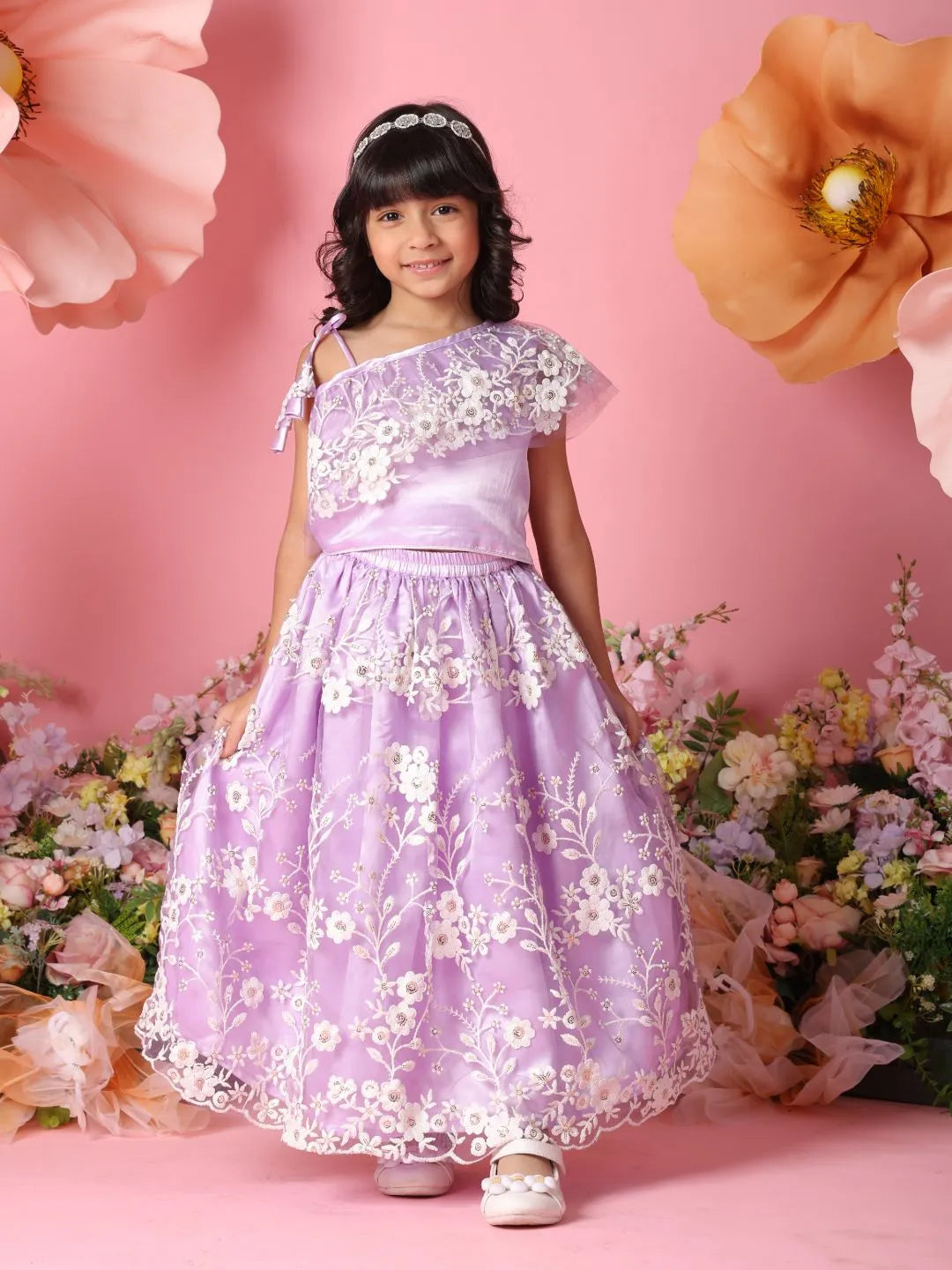 Young girl in a purple floral dress standing against a pink background with flowers.