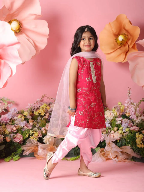 Young girl in a red and pink traditional outfit standing against a pink background with flowers.