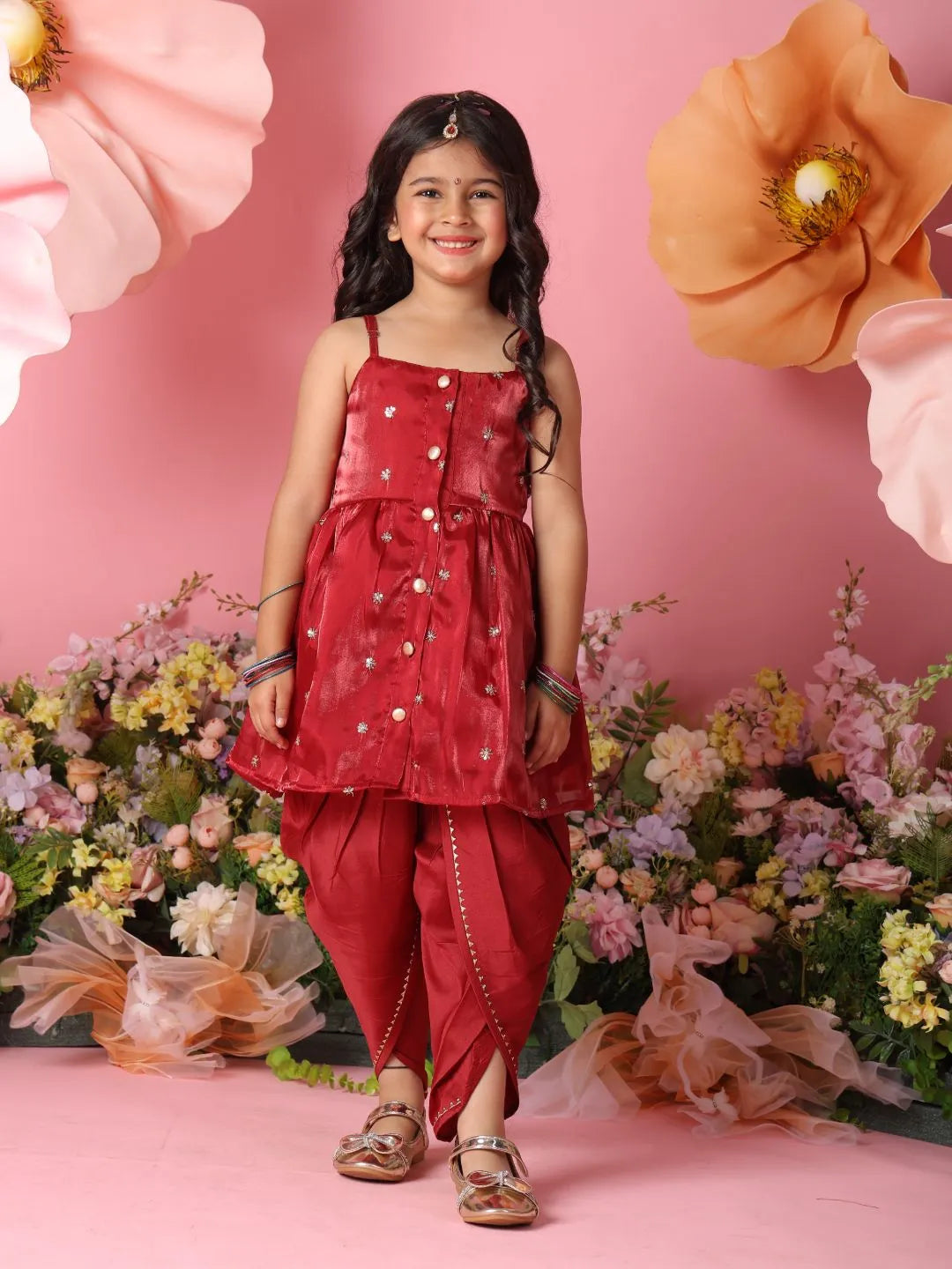 Young girl in a red traditional outfit standing against a pink background with flowers.