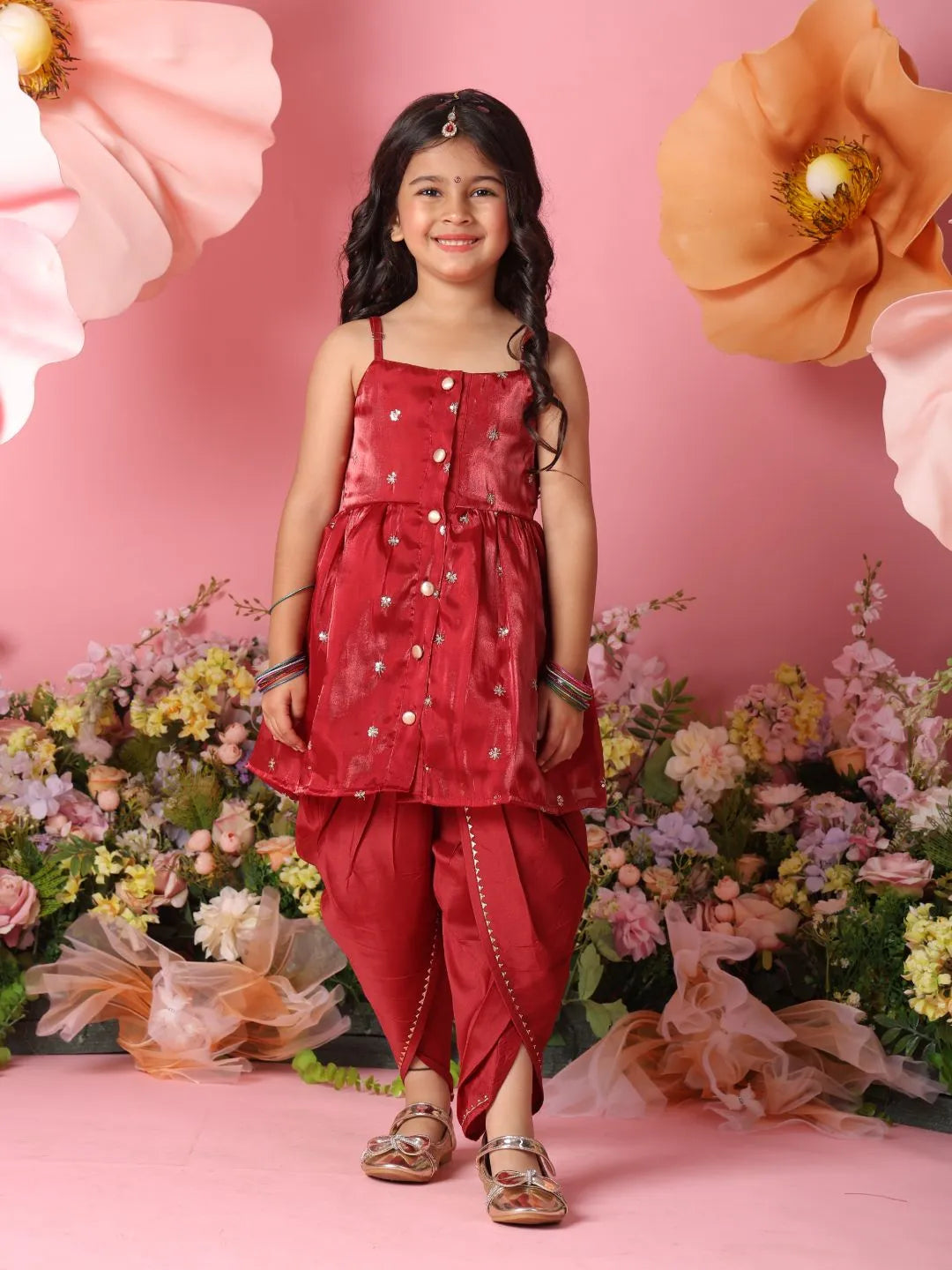 Young girl in a red traditional outfit standing against a pink background with flowers.