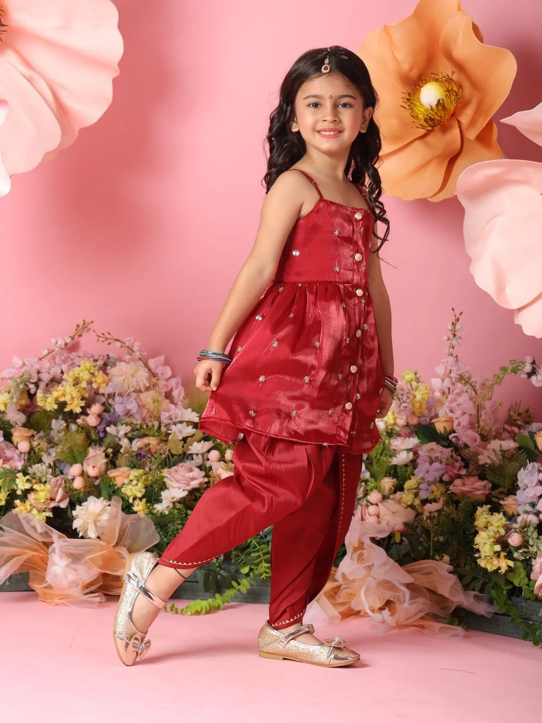 Young girl in a red traditional outfit standing in front of floral decorations on a pink background